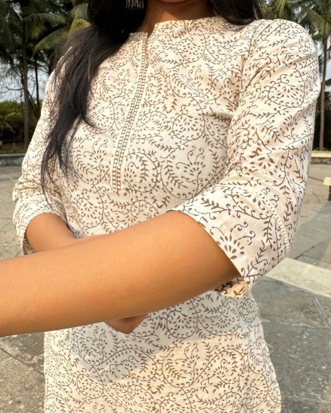 Woman wearing an elegant cream-white cotton kurta set with a light brown plant-inspired print, paired with a matching dupatta. She is posing outdoors against a background of lush green palm trees, exuding a blend of traditional ethnic style and natural charm.